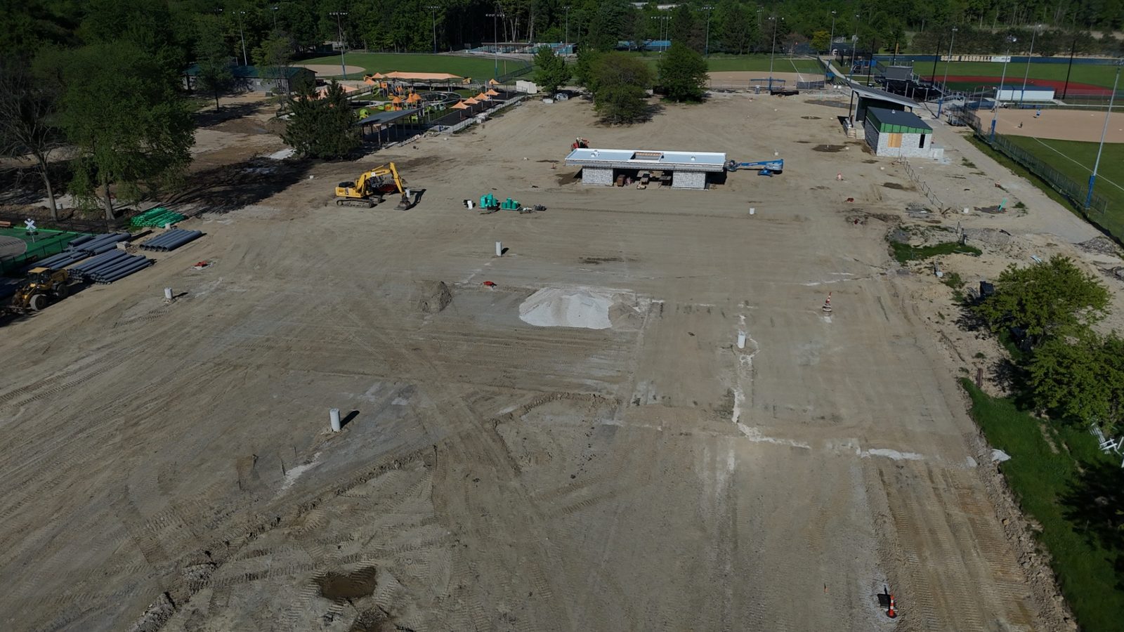 Aerial construction progress photo of a sports complex site under construction — wide overview showing graded fields, an excavator, stockpiled drainage pipe, and a fieldhouse taking shape — Stratum Aerial Media