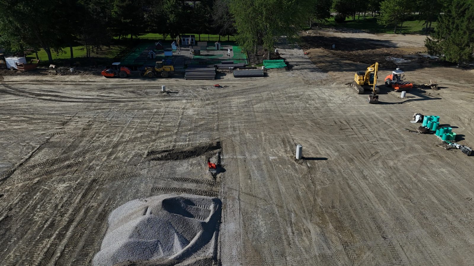 Aerial construction progress photo showing site grading, an excavator, staged drainage materials, and a fieldhouse with a completed roof — Stratum Aerial Media